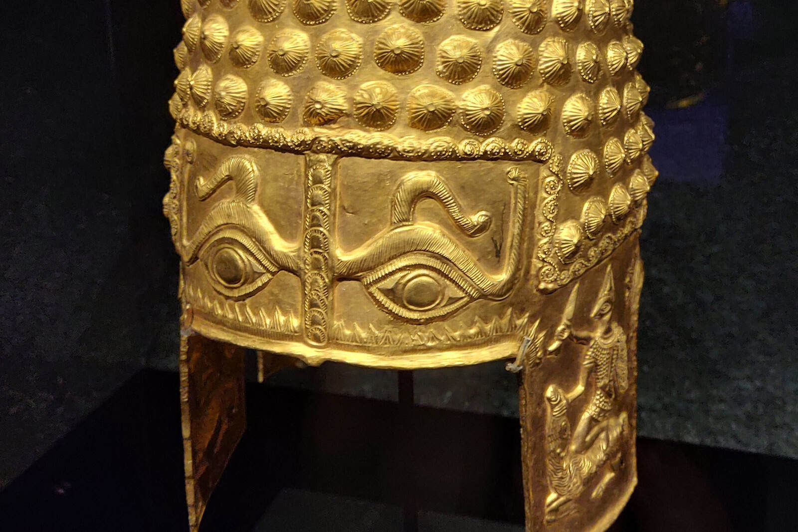 A gold helmet with large embossed eyes and eyebrows, displayed under glass on a dark plinth at the Drents Museum.