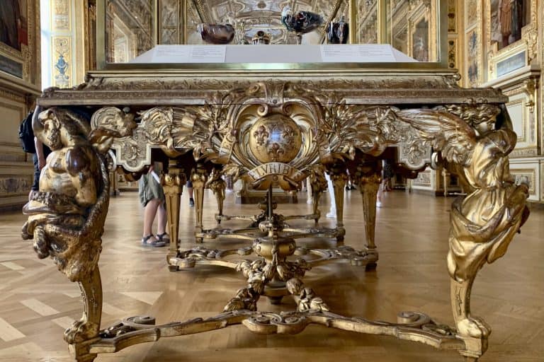 A tall glass display case standing in the Galerie d'Apollon at the Louvre, with the gallery's gilded wall decoration rising behind it.
