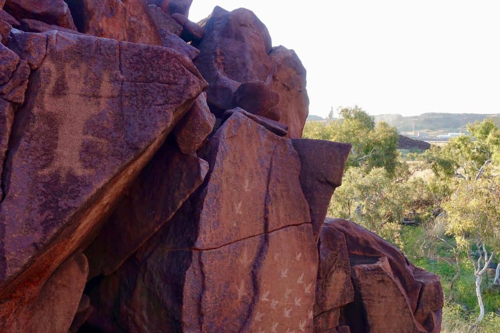 An engraved figure on a dark Murujuga boulder faces a modern ammonia plant across the landscape.