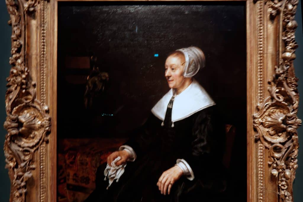 Catrina Hooghsaet sits in a black silk dress and white cap, holding a handkerchief beside a parakeet in Rembrandt’s 1657 portrait.