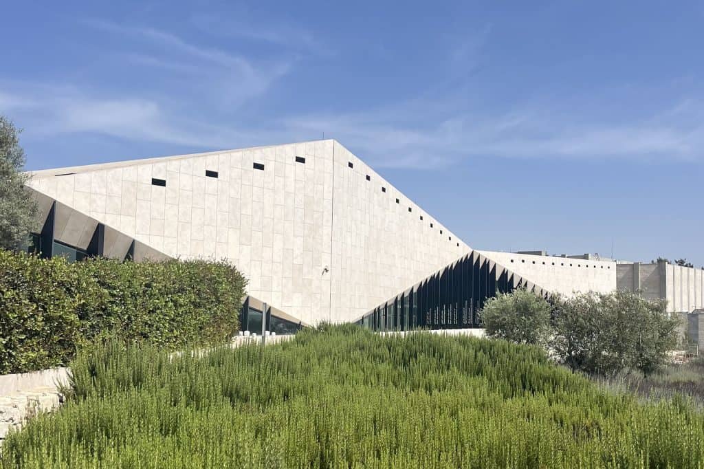 The Palestinian Museum in Birzeit, a low white stone building with angular glass frontage set among terraced hillside gardens under a clear sky.