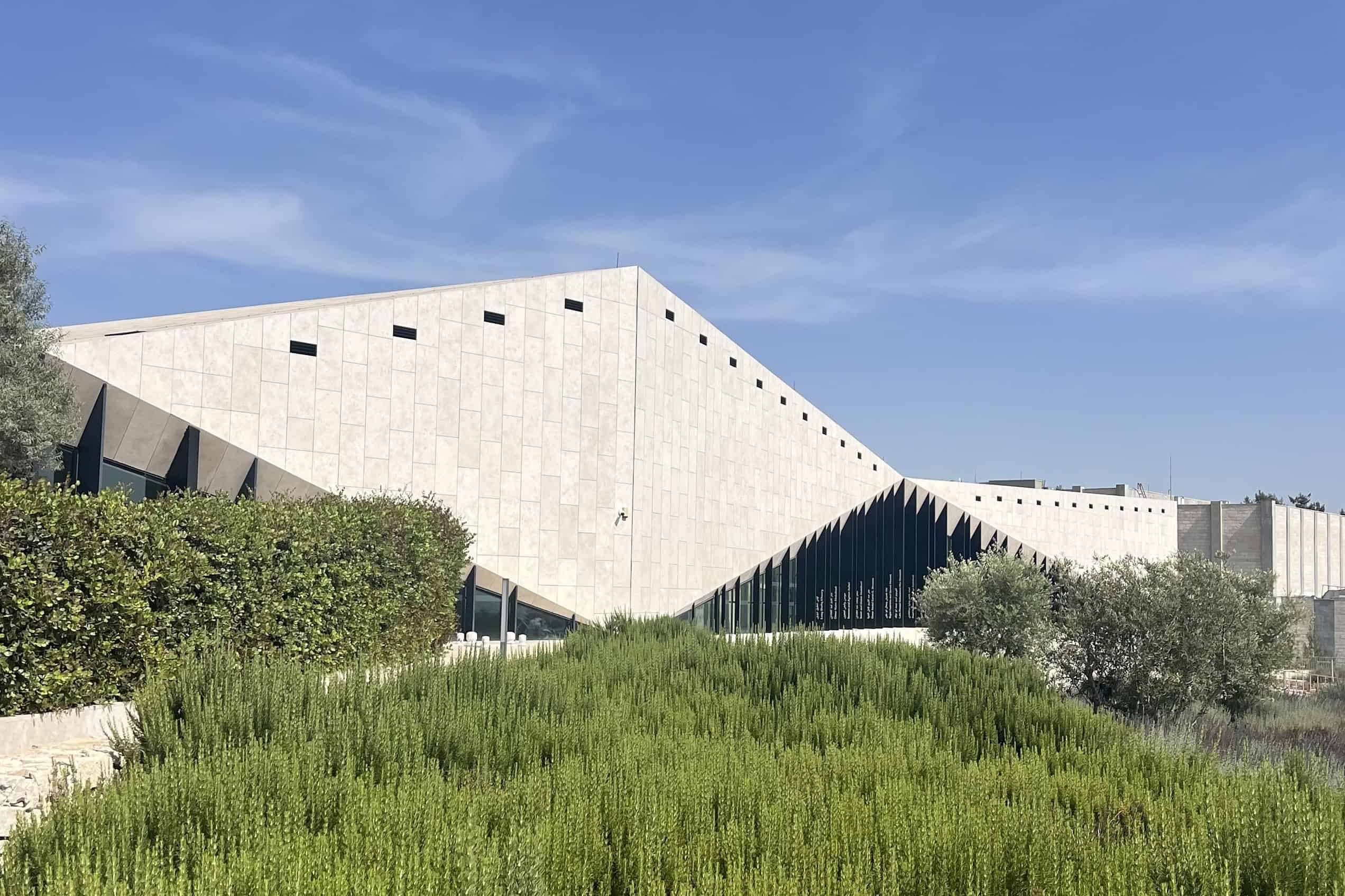 The Palestinian Museum in Birzeit, a low white stone building with angular glass frontage set among terraced hillside gardens under a clear sky.