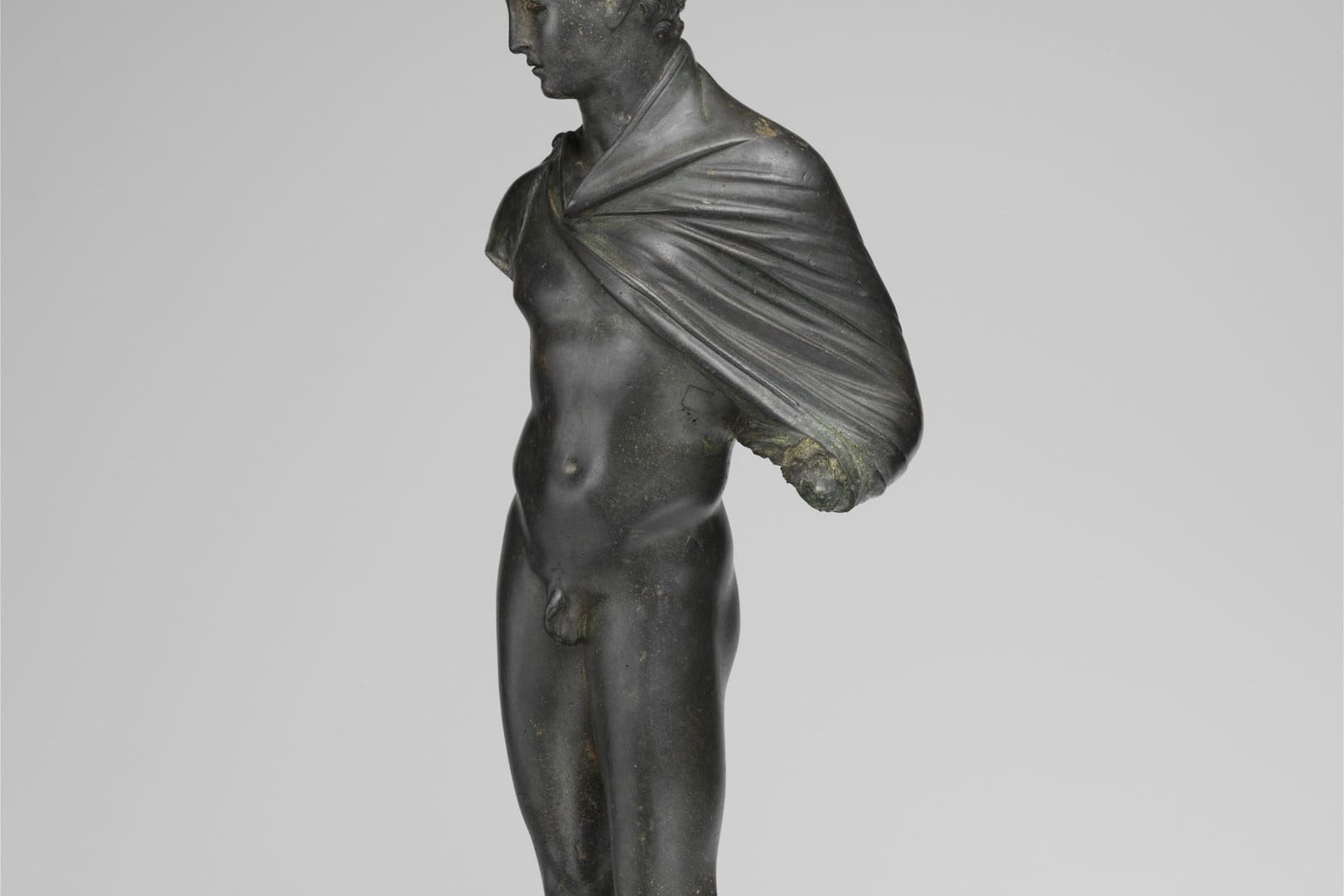 A fragmentary bronze statuette of Hermes stands against a plain background with wings attached at his feet.