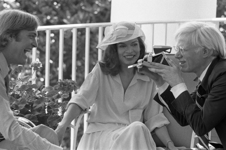 Andy Warhol raises a Polaroid camera while seated between Jack Ford and Bianca Jagger on the Truman Balcony in 1975.