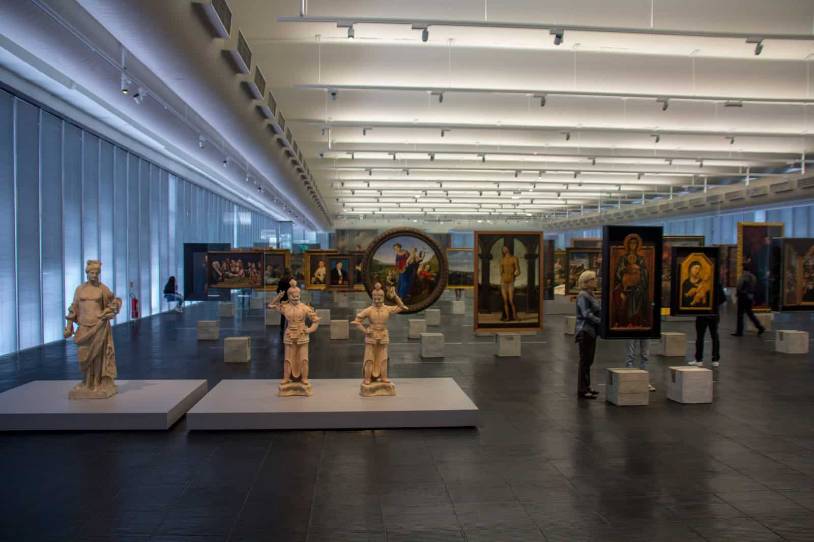 Visitors move among artworks displayed on glass easels inside MASP in São Paulo.