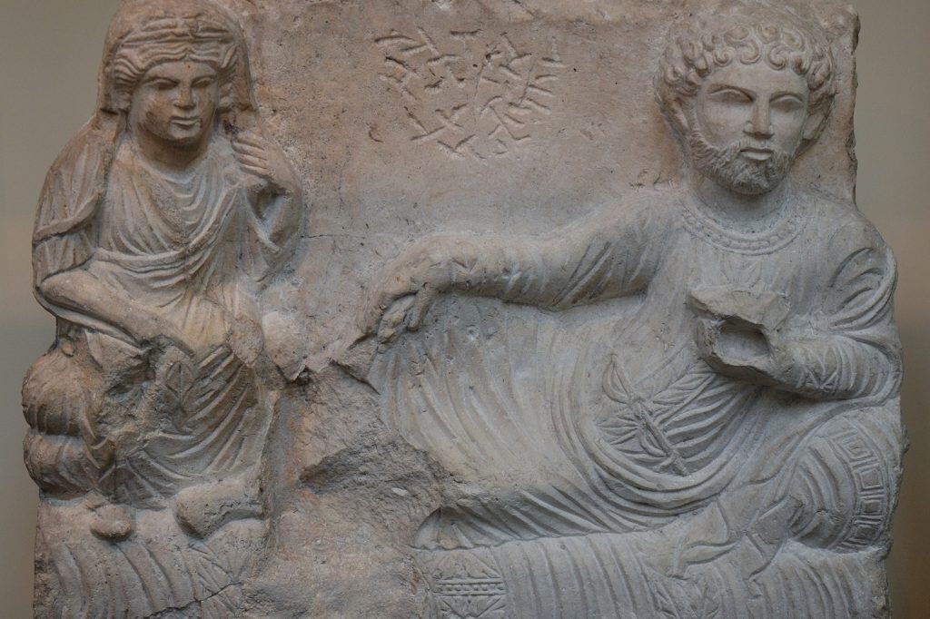 A Palmyrene limestone funerary relief shows a bearded man reclining beside his seated wife beneath a carved inscription.