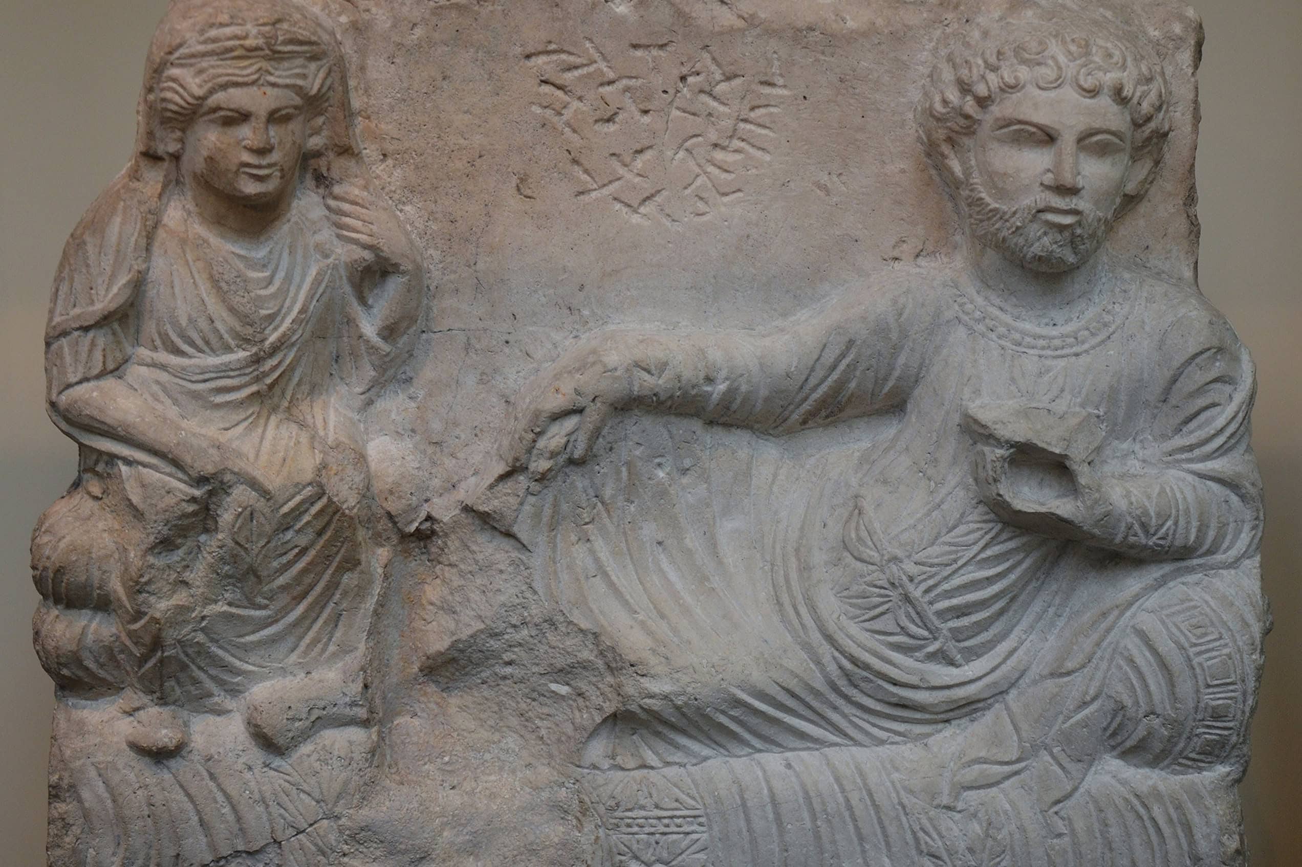 A Palmyrene limestone funerary relief shows a bearded man reclining beside his seated wife beneath a carved inscription.