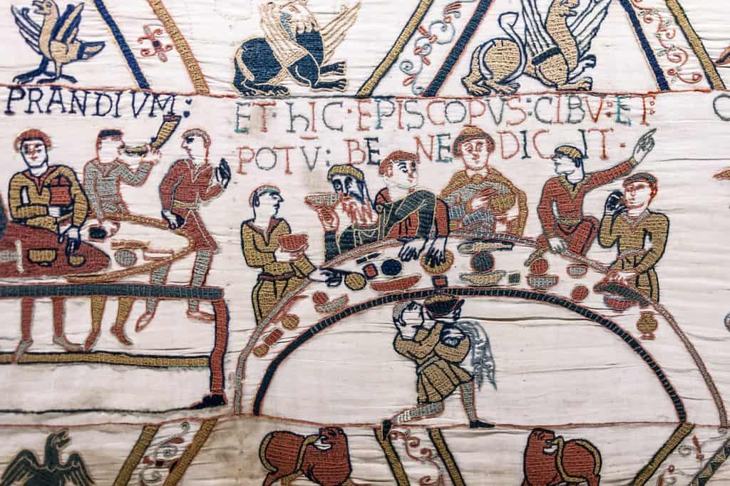 Bishop Odo raises one hand to bless a meal while Norman nobles sit around a curved table in the Bayeux Tapestry.