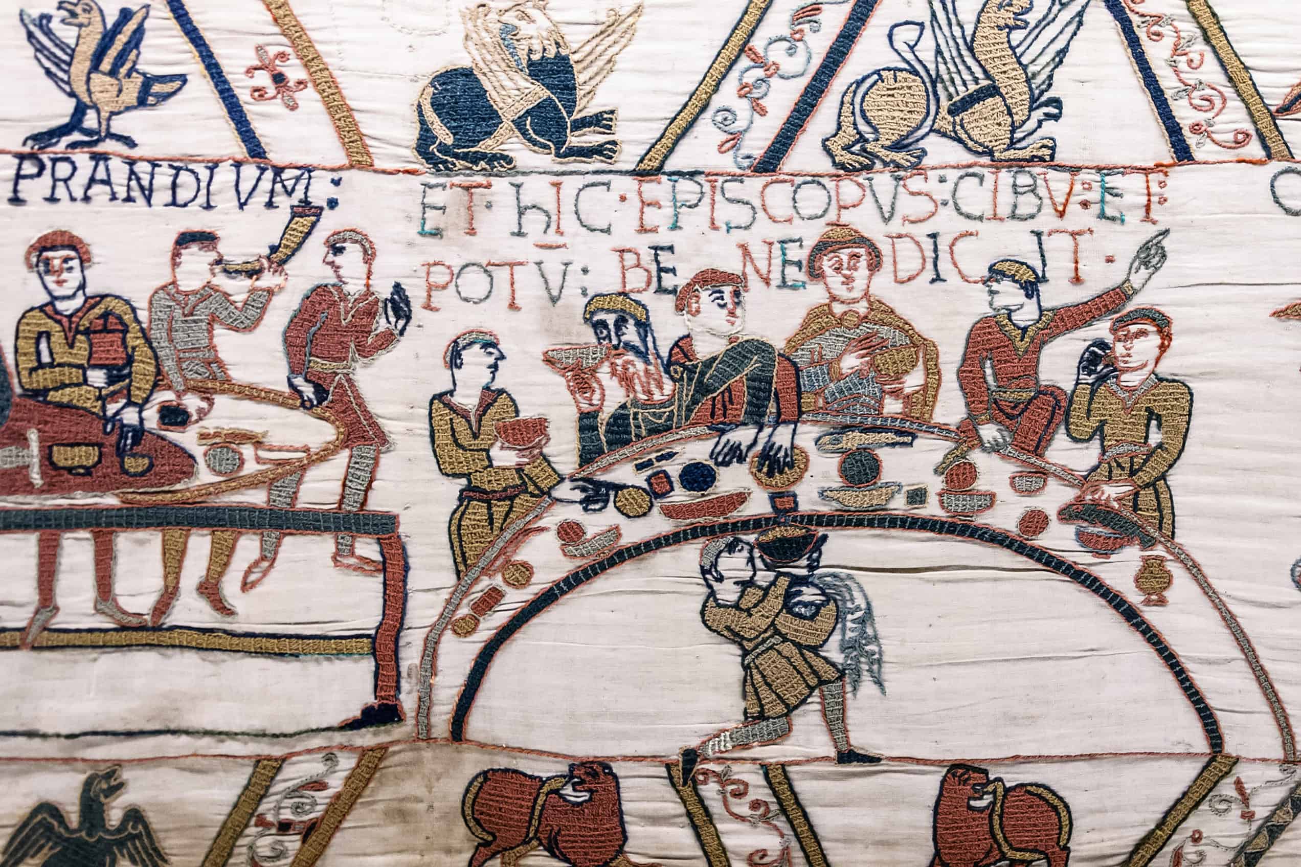 Bishop Odo raises one hand to bless a meal while Norman nobles sit around a curved table in the Bayeux Tapestry.