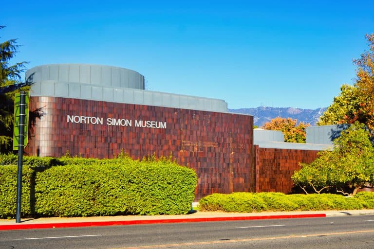 The curved brown façade of the Norton Simon Museum stands behind hedges beneath a clear Pasadena sky.
