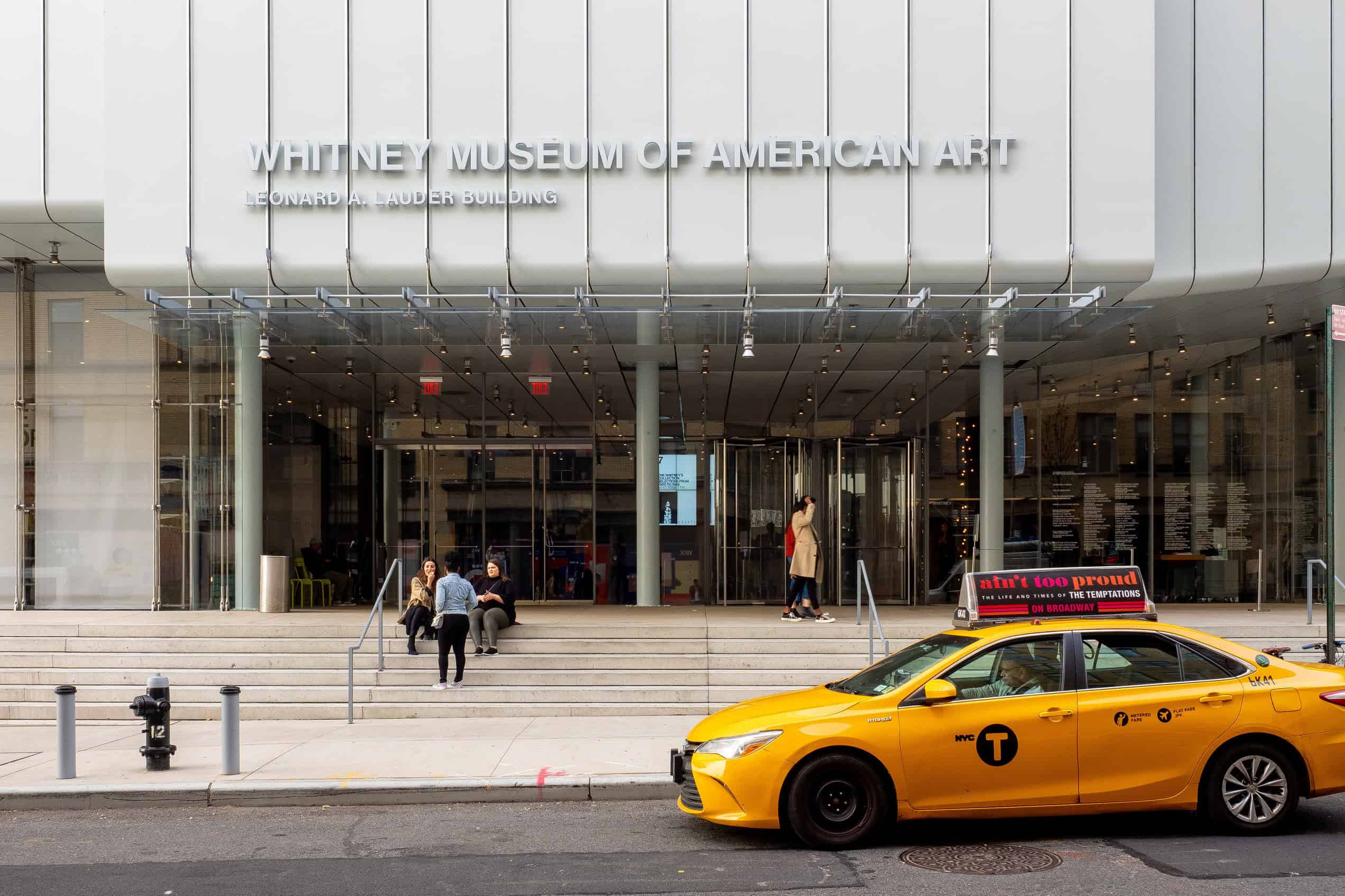 The white-and-glass front of the Whitney Museum rises behind pedestrians and yellow taxis on Gansevoort Street.